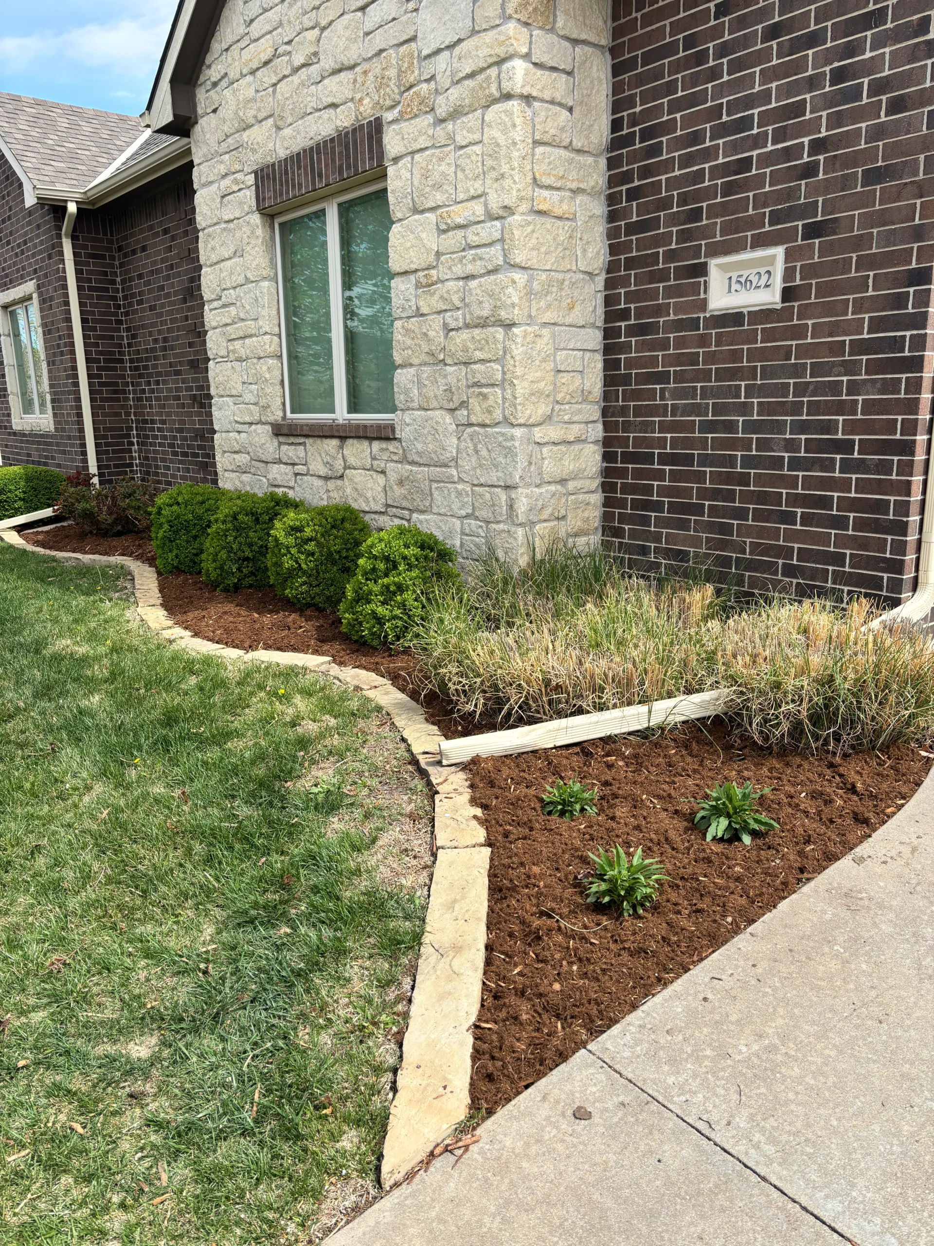 Front yard of a brick-and-stone house with a window, mulch bed, and small shrubs along a curved stone border next to a sidewalk.