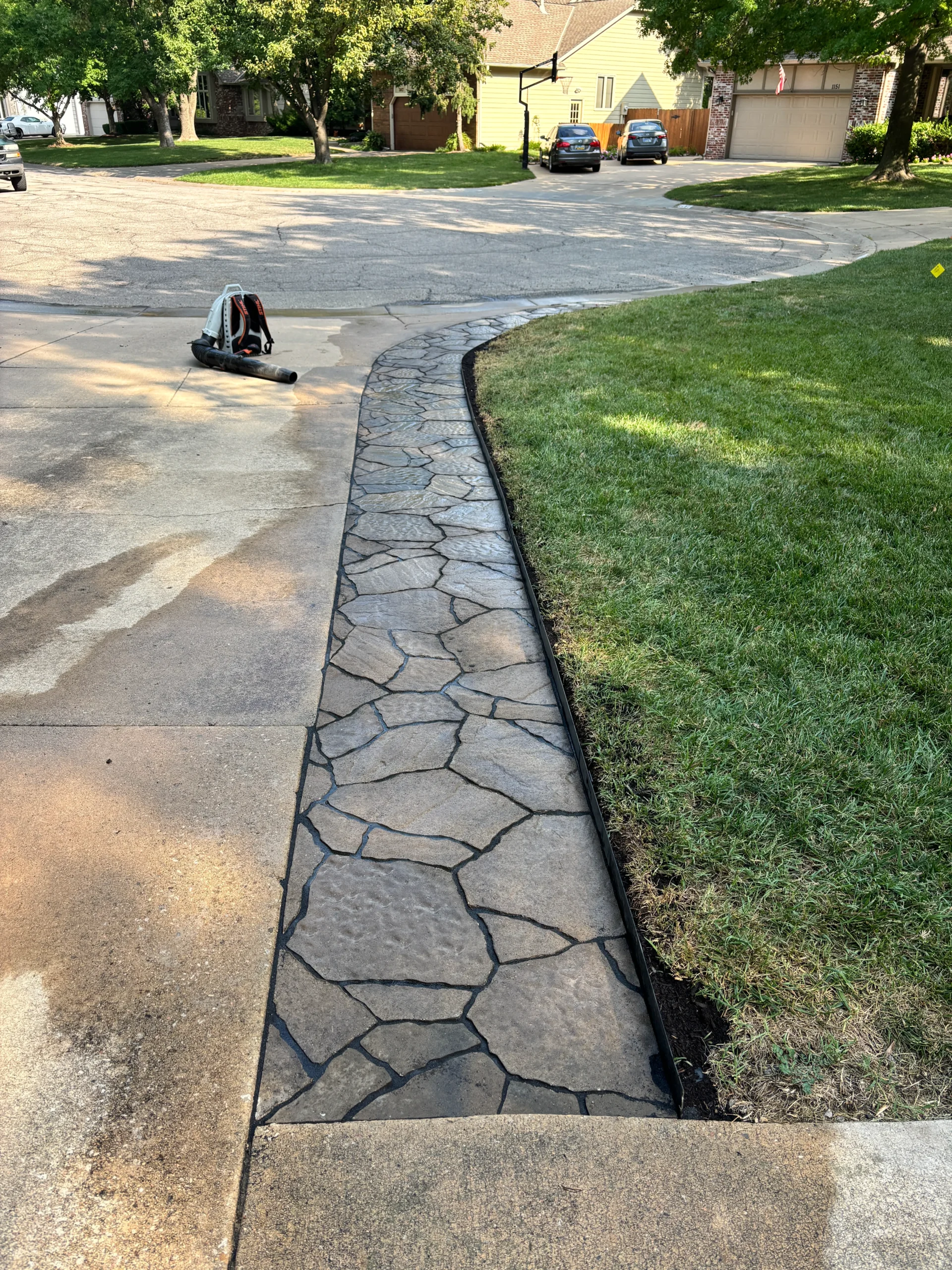 Leaf blower on a concrete driveway beside a curved flagstone edge and a green lawn in a suburban street map area.