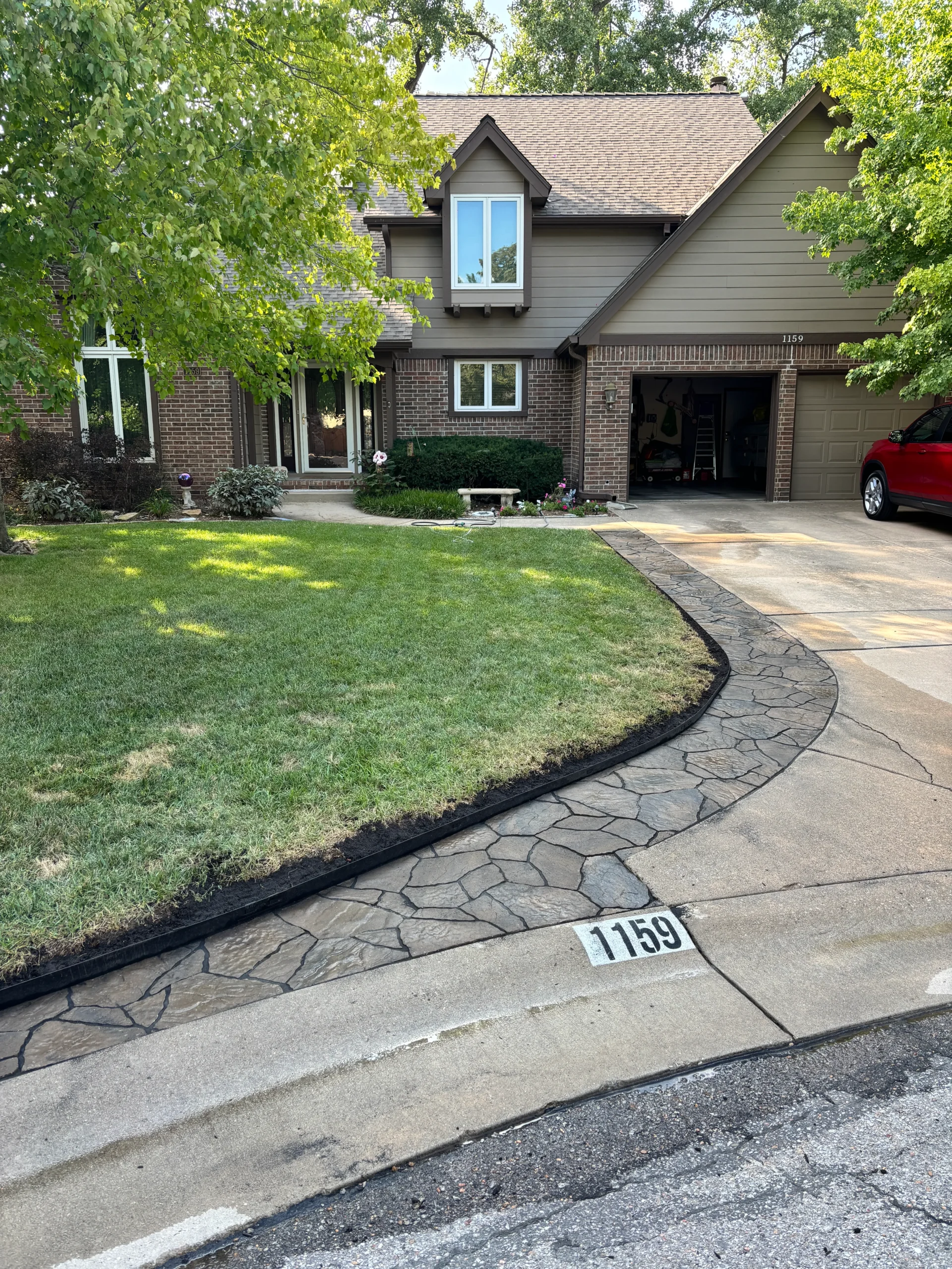 Two-story suburban house with brick and beige siding, a curved stamped concrete driveway, and an open garage; a red car is parked to the right.