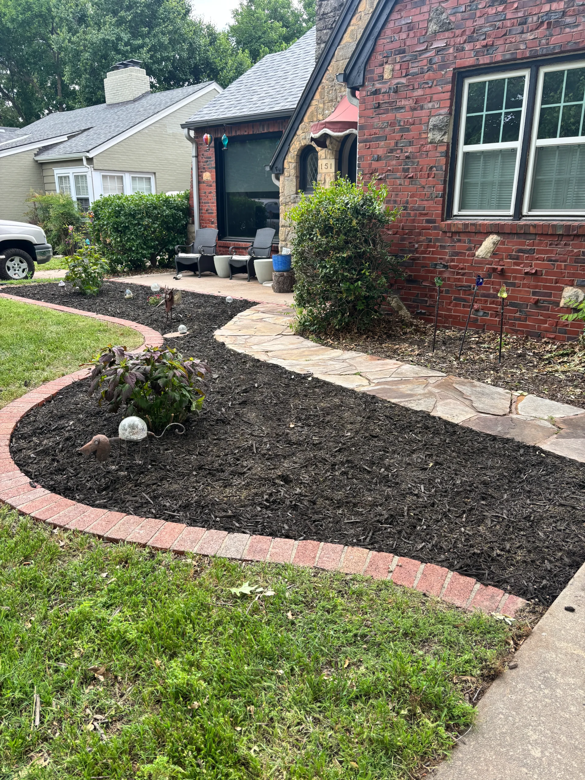 Front yard of a brick house with a curved red-brick border, mulched flower bed, and a stone path leading to a seating area on the porch.