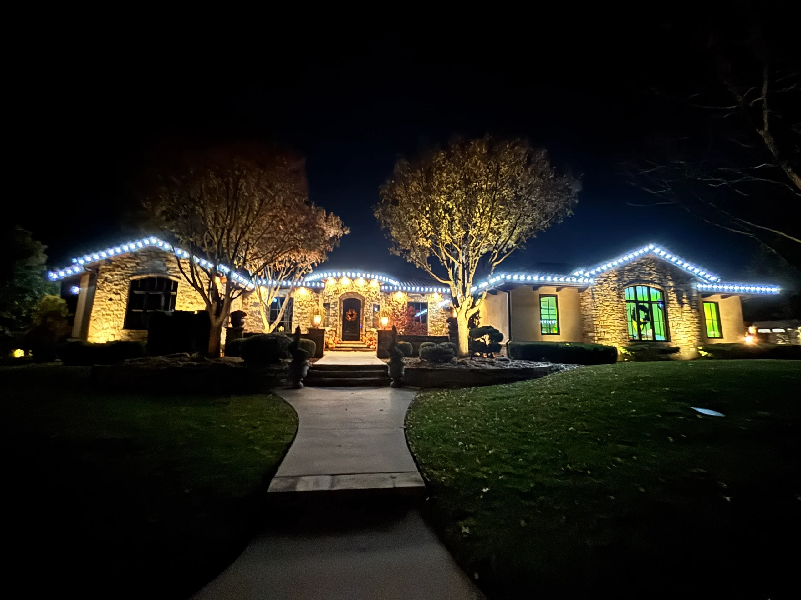 Night scene of a stone house with blue-white string lights along the roof, warm-lit windows, and a curved walkway leading to the entrance.